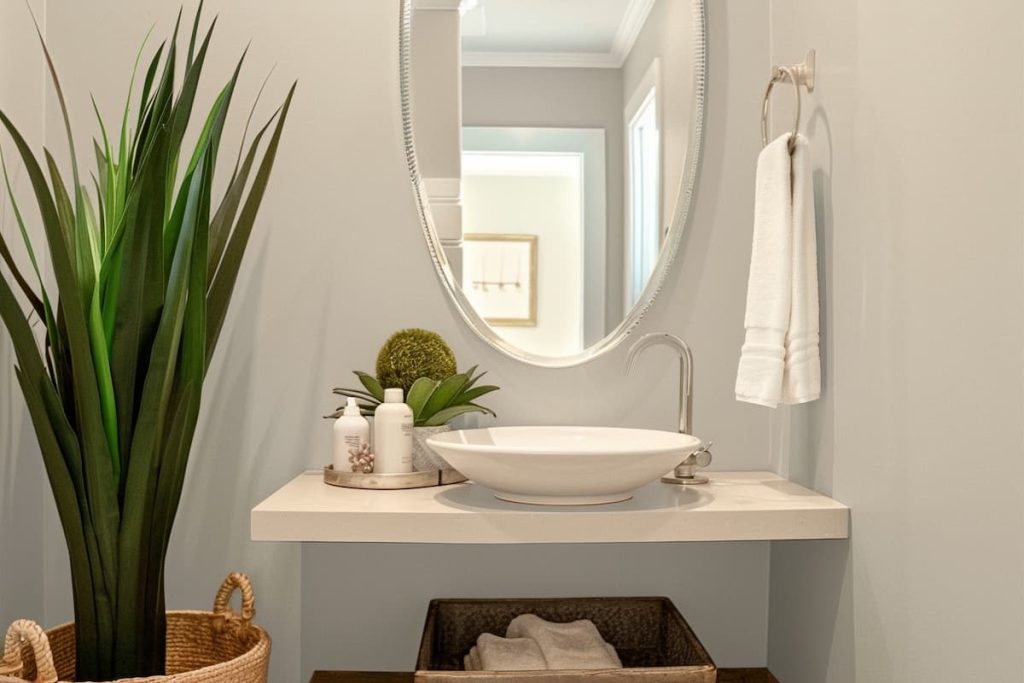 a realistic image of a powder room with a sink, a wall mirror as a face, a large potted plant next to it, toiletries and tissue paper on the shelves. The walls are painted Benjamin Moore Revere Pewter  