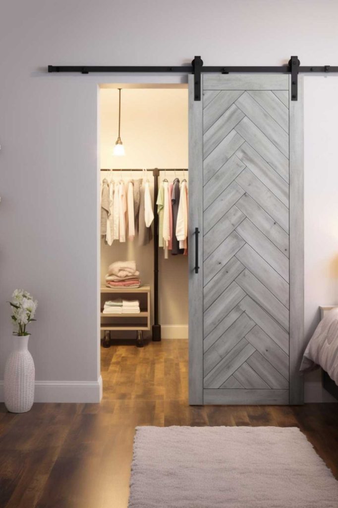 A modern bedroom features a herringbone sliding door that is partially open to reveal a well-organized walk-in closet. On the left there is a white vase with flowers.