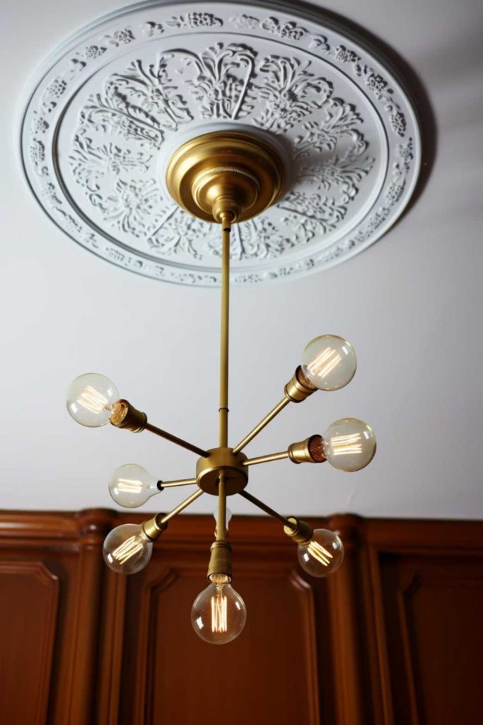 A gold chandelier with eight exposed light bulbs hangs from an ornate white ceiling medallion. Brown paneled walls appear in the background.