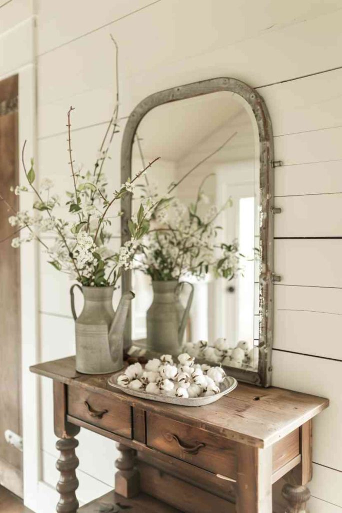 A wooden table with a mirror, two jugs with white flowers and a tray with cotton balls.