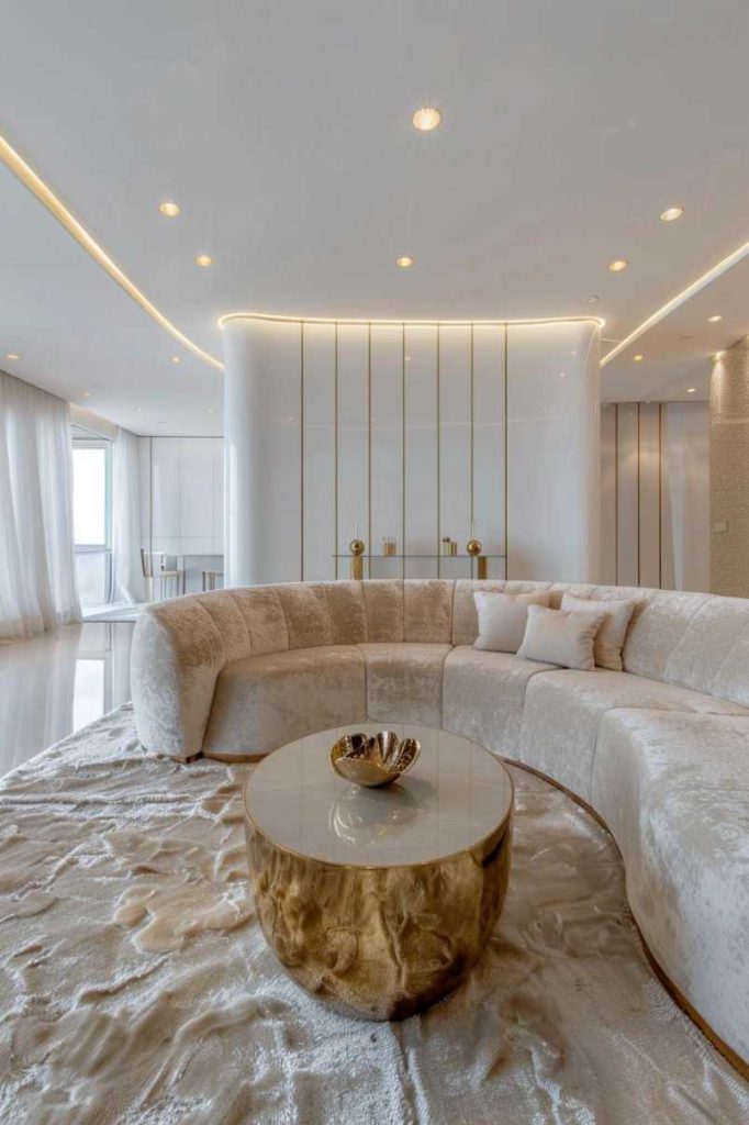 Modern living room with a curved beige sofa, round coffee table with a gold base and minimalist decor. Walls and ceilings have warm lighting accents. Large windows with sheer white curtains.