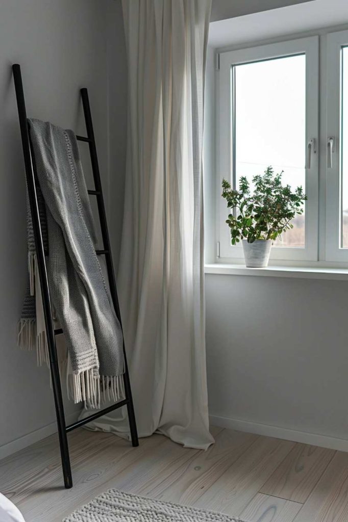 A black ladder holds gray ceilings next to a window with white curtains. A potted plant stands on the windowsill in a minimalist space with light wooden floor.