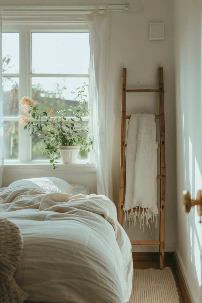 A cozy bedroom with a bed in light bed linen, a window with transparent curtains, a potted plant on the windowsill and a wooden ladder with blankets.