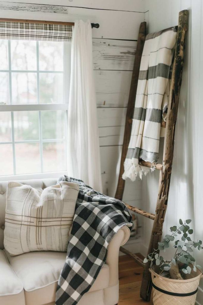 In a cozy corner there is a white sofa with checkered ceilings and a pillow. There are other patterned blankets on a wooden ladder, and there is a potted plant next to a window with white curtains.