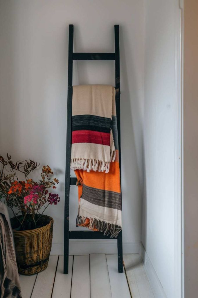 A black ladder in front of a white wall, draped a colorful blanket over it, next to it a basket with flowers.