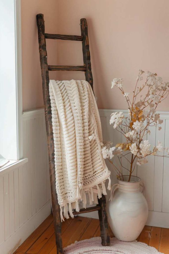 A wooden ladder that leans on a pink wall holds a knitted white ceiling. In addition, there is a white vase with dry flowers on a wooden floor. A window on the left leaves natural light.