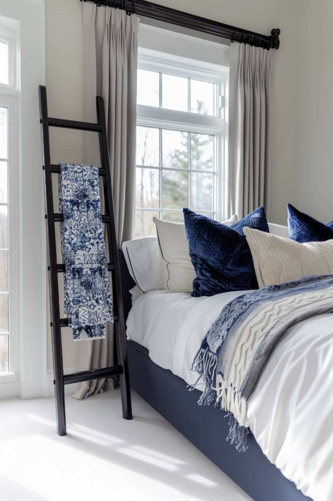 A neatly made bed with blue and white pillows and ceilings stands next to a black ladder with blue patterned fabric in a room with large windows and white curtains.