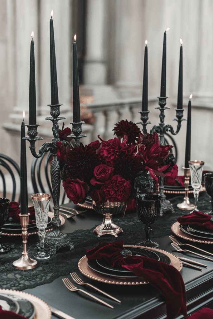 Elegant table setting with black candles in ornate candle holders, dark red flowers, gold-rimmed plates and black and gold cutlery on a black lace tablecloth.