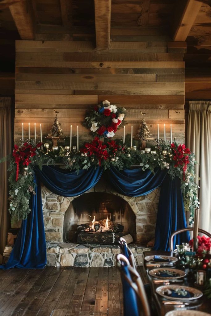 A fireplace with deep blue velvet, green garlands and red flower arrangements. The fireplace contains numerous candles and a festive wreath adorns the wall above. A wooden table with space settings is visible to the side.