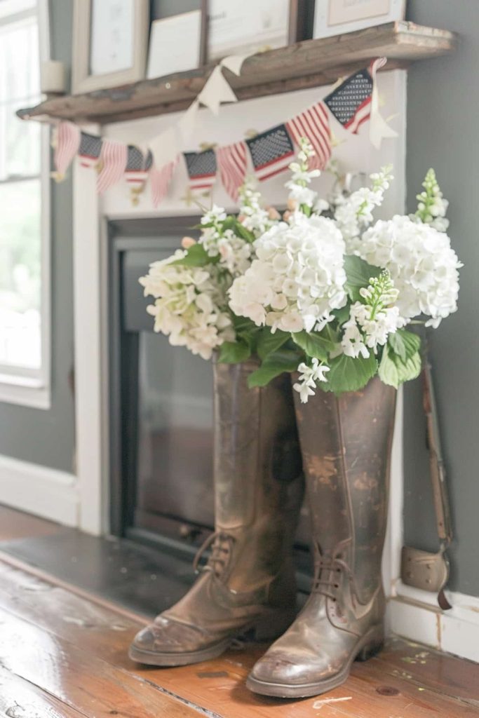 Old leather boots with white hydrangea flowers are placed in front of a fireplace. At the top contains a rustic shelf framed pictures and American flag-bullet.