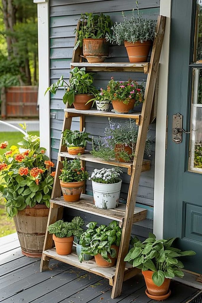 A wooden conductor shelf against a gray house wall filled with different potted plants such as herbs and flowers. There is a blue door nearby and a large pot with living orange flowers on the deck. 