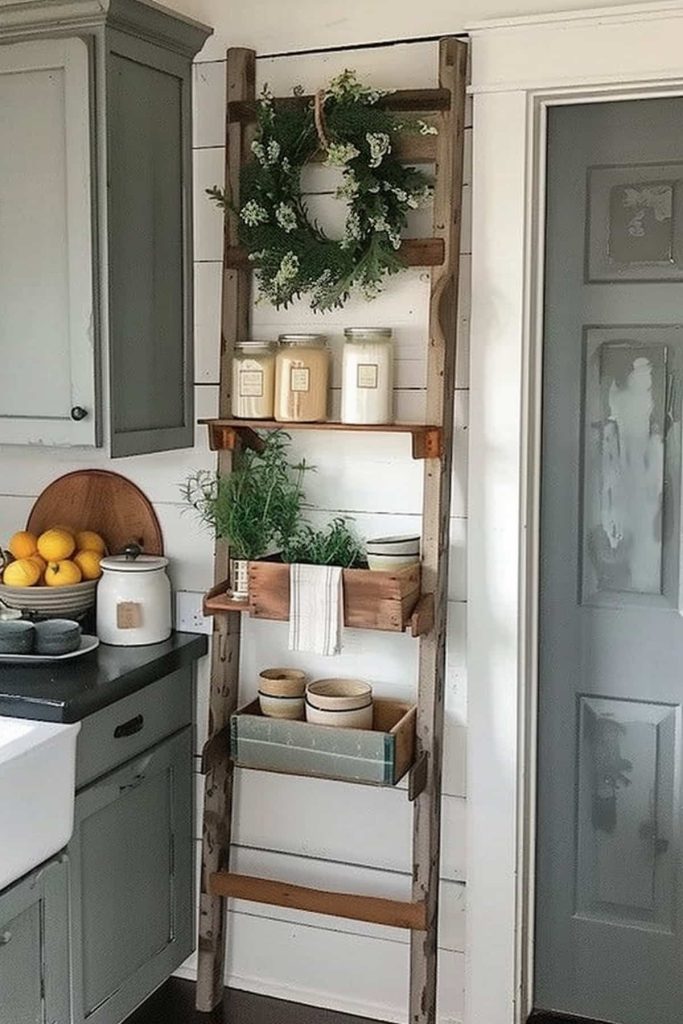 A rustic wooden ladder is leaned against a white Shipap wall in a kitchen, which serves as a shelf. The shelves hold candles, a small potted plant and bowls. A wreath with green hangs at the top. There is a counter with a bowl of lemons on the left.