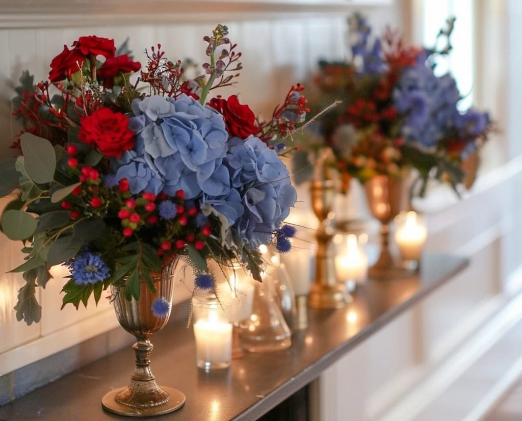 A fireplace that is adorned with elegant flower arrangements with red roses, blue hydrangeas and various green reasons. Candles in brass owners and glass votives are illuminated